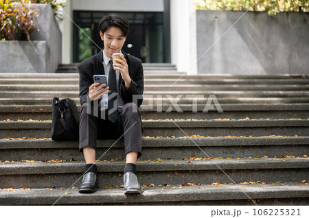 A happy Asian businessman is using his smartphone and sipping coffee while sitting on a staircase A happy Asian businessman is using his smartphone and sipping coffee while sitting on a staircase 106225321