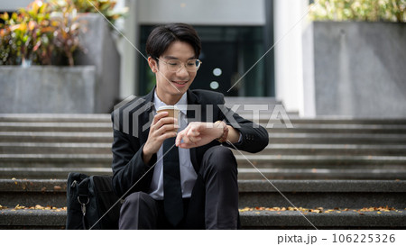 A smart businessman is sipping coffee and checking time on his watch while sitting on a staircase A smart businessman is sipping coffee and checking time on his watch while sitting on a staircase 106225326