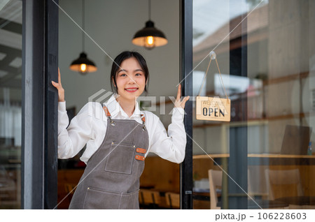 A cheerful Asian female coffee shop owner or waitress stands at the entrance door of her shop. 106228603