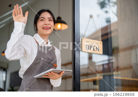 A pretty Asian female coffee shop worker stands at the entrance door with a tablet in her hand 106228608