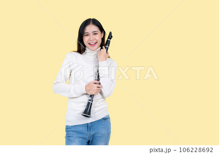 An attractive Asian female musician with a clarinet stands against an isolated yellow background. 106228692