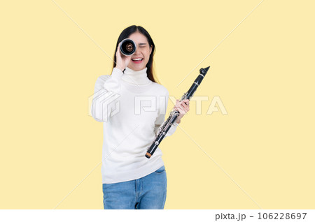 A playful Asian female musician with a clarinet stands against an isolated yellow background. 106228697