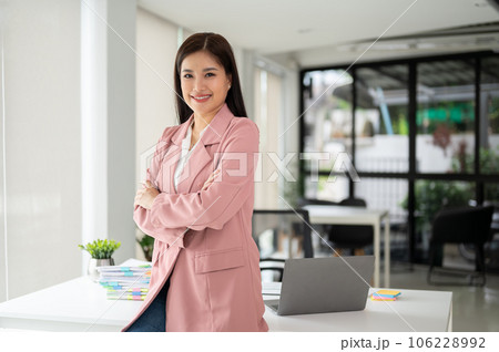 A beautiful Asian businesswoman stands in the office with her arms crossed, smiling at the camera. 106228992