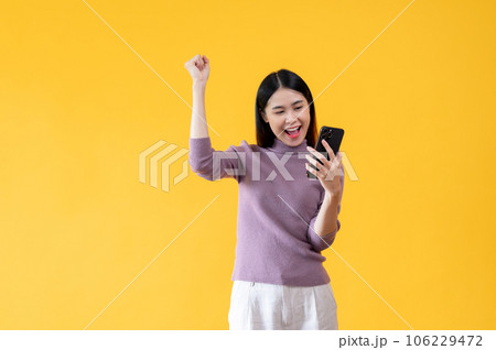 A cheerful Asian woman is showing clenched fist and looking at her smartphone with a happy face A cheerful Asian woman is showing clenched fist and looking at her smartphone with a happy face 106229472