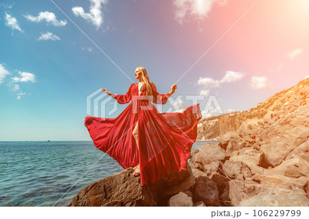 Red dress sea woman. A blonde with flowing hair in a long flowing red dress stands on a rock near the sea. Travel concept, photo session at sea Red dress sea woman. A blonde with flowing hair in a long flowing red dress stands on a rock near the sea. Travel concept, photo session at sea 106229799