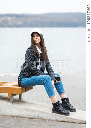 A young pretty girl, dressed in a leather jacket and jeans, spends time alone on the river. Girl holding a mobile phone in his hands 106237460