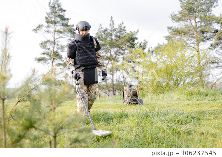 A man in a military uniform and bulletproof vest works in the forest with a metal detector. A minesweeper performs work on demining the territory 106237545