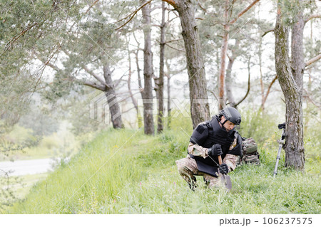 A man in a military uniform and a bulletproof vest works in the forest to demine the territory. A man excavates explosives in the forest A man in a military uniform and a bulletproof vest works in the forest to demine the territory. A man excavates explosives in the forest 106237575