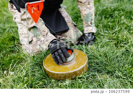 A man in a military uniform and a bulletproof vest works in the forest to demine the territory. A man warns of danger by making a red mark 106237599
