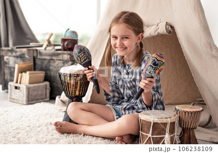 Smiling little girl shaking music maracas sitting in room with djembe drums and wigwam Smiling little girl shaking music maracas sitting in room with djembe drums and wigwam 106238109