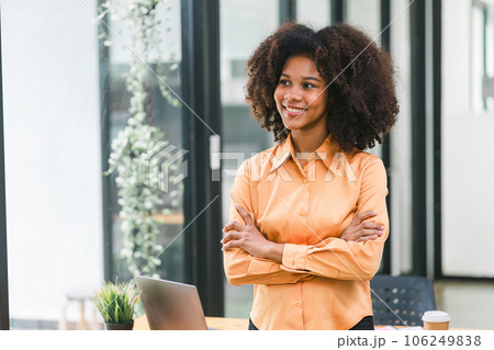 A beautiful young African American woman, smiling at the camera while standing with her arms crossed in a modern office. 106249838