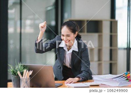 Portrait of a happy young businesswoman celebrating success with arms raised in front of a laptop, fists clenched. The freelancer has finished a project. 106249900