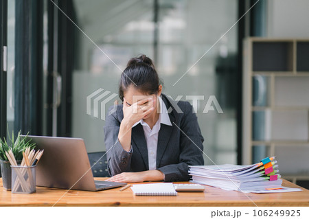 The stressed and exhausted millennial Asian businesswoman is seen sitting at her office desk with her hand on her head, indicating a hard working day where she is overloaded with work. 106249925
