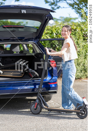 旅行先の駐車場で電動キックボードを車のトランクに収納する若くて可愛い女性 106249958