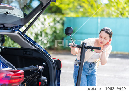 旅行先の駐車場で電動キックボードを車のトランクにどのように収納するか悩む若くて可愛い女性 106249960