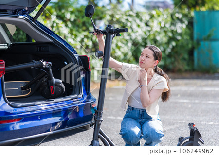 旅行先の駐車場で電動キックボードを車のトランクにどのように収納するか悩む若くて可愛い女性 106249962