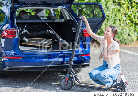 旅行先の駐車場で電動キックボードを車のトランクにどのように収納するか悩む若くて可愛い女性 106249965