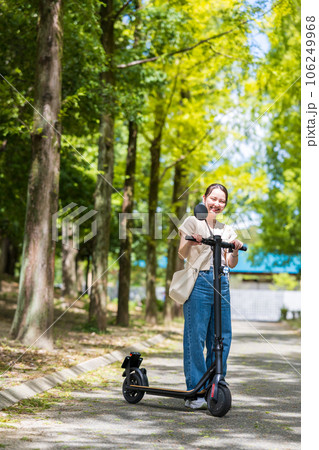 緑が美しい並木道を電動キックボードで爽快に走る若くてかわいい女性｜電動キックボードイメージ 106249968