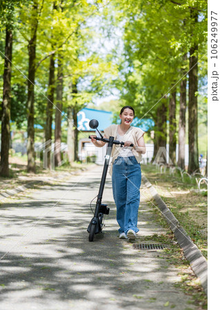 緑が美しい並木道を電動キックボードで爽快に走る若くてかわいい女性｜電動キックボードイメージ 106249977
