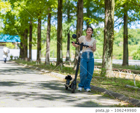 緑が美しい並木道を電動キックボードで爽快に走る若くてかわいい女性｜電動キックボードイメージ 106249980