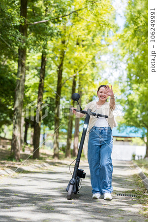 緑が美しい並木道に電動キックボードで訪れ手を振る若くてかわいい女性｜電動キックボードイメージ 106249991