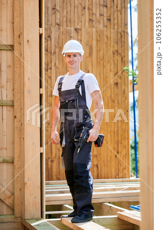 Portrait of carpenter constructing wooden framed house. Man worker cladding facade of house, using screwdriver. Concept of modern eco-friendly construction. 106255352