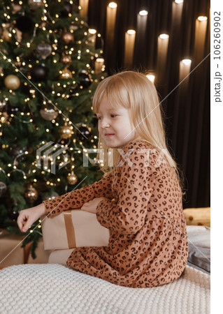 A little blonde girl is sitting on a soft bed in a festive interior decorated in a New Year's style. A child holds a Christmas gift in a craft package. 106260902