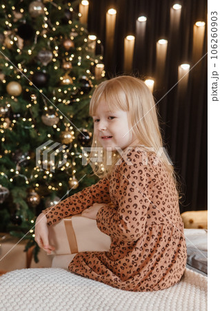 A little blonde girl is sitting on a soft bed in a festive interior decorated in a New Year's style. A child holds a Christmas gift in a craft package. 106260903
