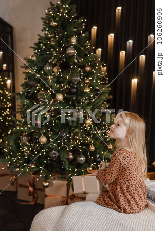 A little blonde girl is sitting on a soft bed in a festive interior decorated in a New Year's style. A child holds a Christmas gift in a craft package. 106260906