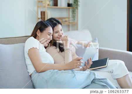 lgbtq, lgbt concept, homosexuality, portrait of two asian women posing happy together and loving each other while playing tablet at sofa. lgbtq, lgbt concept, homosexuality, portrait of two asian women posing happy together and loving each other while playing tablet at sofa. 106263377
