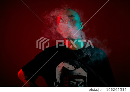 Close-up studio shot of a young bald guy vaping, blowing out a cloud of smoke on red background. Close-up studio shot of a young bald guy vaping, blowing out a cloud of smoke on red background. 106265555