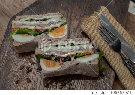 Sandwich stuffed with chicken breast, cheese, boiled eggs and fresh vegetables serve with fork and knife on wooden plate. 106267973
