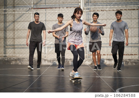 young asian woman skateboarder skateboarding outdoors with friends watching from behind young asian woman skateboarder skateboarding outdoors with friends watching from behind 106284588