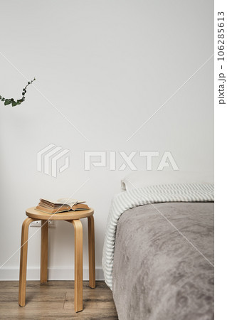 Close-up of the bedroom interior, a headboard with light-colored linens, a stool instead of a bedside table, there is an open book on it. Home interior in a minimalist style 106285613