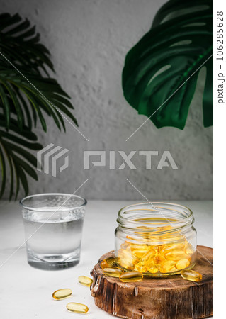 Close-up on capsules of fish oil with omega-3 and vitamin D in a glass jar on a wooden stand, a glass of water next to it, health care concept. Fish Oil Softgels Close-up on capsules of fish oil with omega-3 and vitamin D in a glass jar on a wooden stand, a glass of water next to it, health care concept. Fish Oil Softgels 106285628