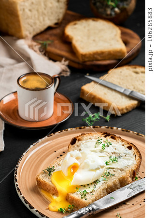 Soft-boiled (poached) egg on slice of bread covered with butter cream and herbs, on clay plate on black wooden table. Espresso coffee and loaf of sliced bread on blurred background. Breakfast idea Soft-boiled (poached) egg on slice of bread covered with butter cream and herbs, on clay plate on black wooden table. Espresso coffee and loaf of sliced bread on blurred background. Breakfast idea 106285883