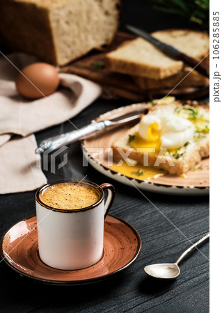 Close-up of cup of espresso coffee with rising steam on black wooden table. On blurred background, soft-boiled egg (poached) in slice of bread, with butter cream and herbs. Breakfast idea Close-up of cup of espresso coffee with rising steam on black wooden table. On blurred background, soft-boiled egg (poached) in slice of bread, with butter cream and herbs. Breakfast idea 106285885