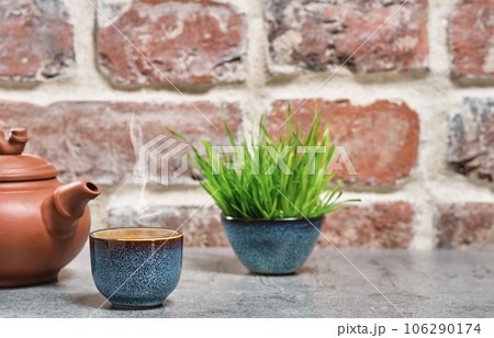 Hot green tea in a blue bowl, selective focus, steam rises above the cup, next to a clay teapot. Gray stone table, brick vintage background. Close-up, tea ceremony, minimalism, place for text 106290174
