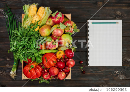 Vegetables and fruits and herbs in a box layout on a dark wooden table, next to a notebook and a pencil for keeping track of the diet. Fresh food delivery from farmers market, seasonal food Vegetables and fruits and herbs in a box layout on a dark wooden table, next to a notebook and a pencil for keeping track of the diet. Fresh food delivery from farmers market, seasonal food 106290368