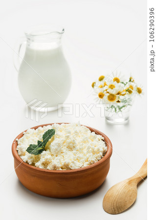 Cottage cheese in a ceramic rustic cup, next to a wooden spoon and a jug of milk. Close-up, selective focus, bright white background. Soft curd , natural healthy food, complete diet food 106290396