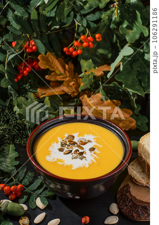 Vegetarian pumpkin soup in black bowl with cream and pumpkin seeds, Close-up, selective focus on soup and seeds. On a dark wooden table, plate of soup stands surrounded by autumn oak and rowan leaves Vegetarian pumpkin soup in black bowl with cream and pumpkin seeds, Close-up, selective focus on soup and seeds. On a dark wooden table, plate of soup stands surrounded by autumn oak and rowan leaves 106291186