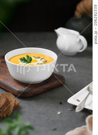 Pumpkin chowder with cream, pumpkin seeds and parsley in a white bowl on a gray background. Seasonal vegetarian soup, selective focus on parsley Pumpkin chowder with cream, pumpkin seeds and parsley in a white bowl on a gray background. Seasonal vegetarian soup, selective focus on parsley 106292339