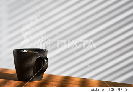 Cup of freshly made espresso coffee in a cup on a wooden table, steam rises over the ristretto. Coffee background. The wall of the cafe is covered with sunny stripes from the blinds. Coffee break Cup of freshly made espresso coffee in a cup on a wooden table, steam rises over the ristretto. Coffee background. The wall of the cafe is covered with sunny stripes from the blinds. Coffee break 106292350