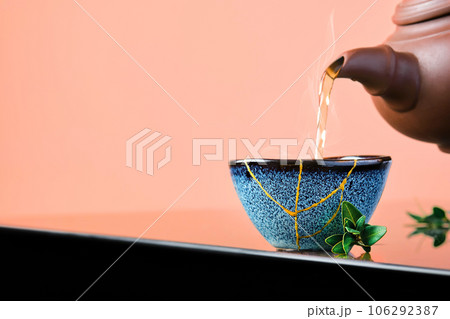 Hot green tea is poured from a ceramic teapot into a bowl. Selective focus on the blue cup. Steam will rise above the mug Reclaimed ceramic blue cup, second life of things, recycling or kintsugi 106292387