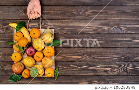 Various ripe, raw fruits in package. Woman's hand holds package, dark restored wooden background. Top view. Food paper bag idea of zero waste. Food delivery, purchase or donation concept. Copy space 106293949