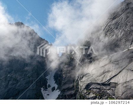 Mountain pass in the clouds. Gloomy minimalist foggy mountain landscape with snow cornice. Dangerous mountain pass above precipice. Danger rocks in thick fog. Mountain pass in the clouds. Gloomy minimalist foggy mountain landscape with snow cornice. Dangerous mountain pass above precipice. Danger rocks in thick fog. 106297214