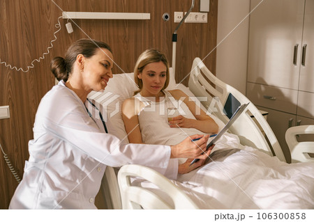Doctor visiting woman patient in hospital ward to check his health state and looking at camera 106300858