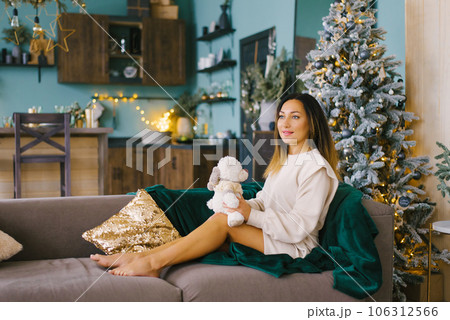 Pregnant woman holds a soft toy in her hands and rests on the sofa near the Christmas tree Pregnant woman holds a soft toy in her hands and rests on the sofa near the Christmas tree 106312566