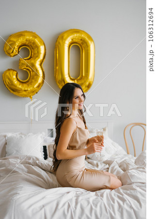 Long haired birthday woman sitting on the bed in the morning and holds a glass of champagne in her hands and smiles celebrating her thirtieth birthday 106312694