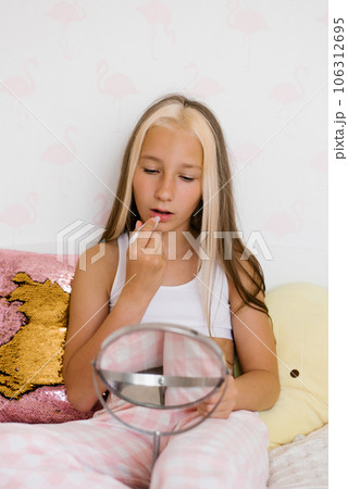 Caucasian teenage girl is sitting on the bed, looking in the mirror and painting her lips with balm 106312695